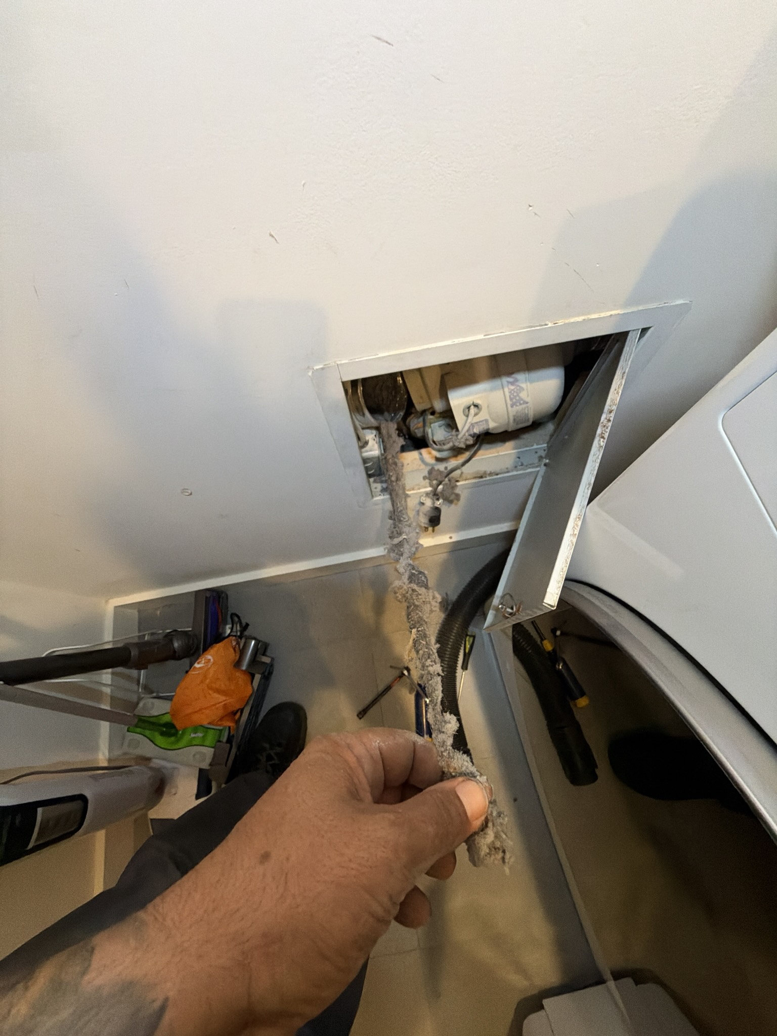 Open access panel behind a dryer showing semi-rigid metal duct heavily coated in matted lint, with a gloved hand pulling out a compacted lint clump beside a booster fan and electrical outlet