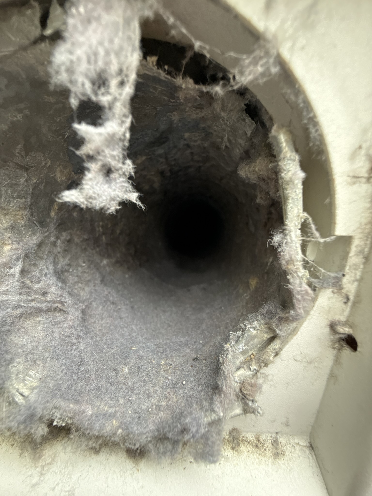 Interior view of a dryer vent duct opening severely clogged with compacted lint, leaving only a small dark passage through the center of the duct